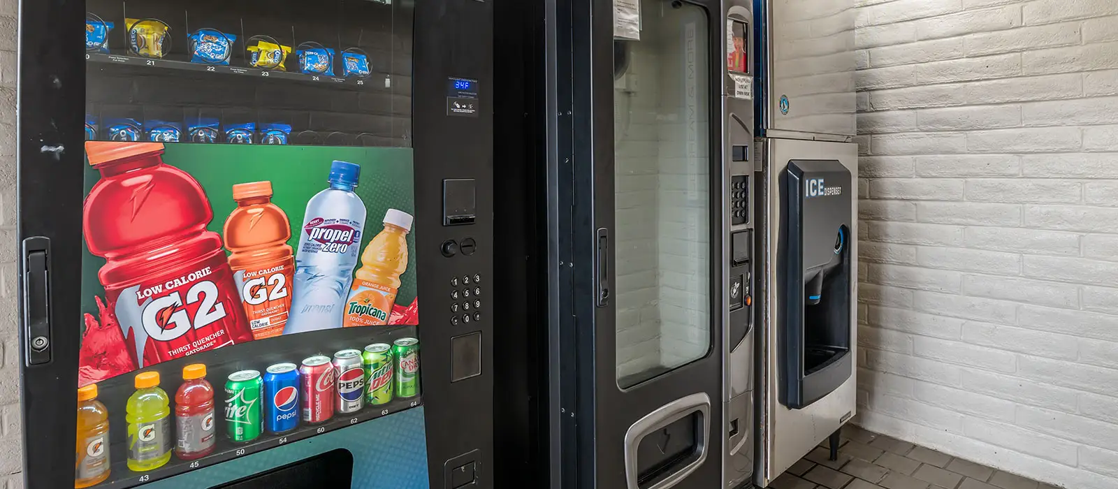 A vending machine with drinks and snacks, a second vending machine, and an ice dispenser are against a white brick wall