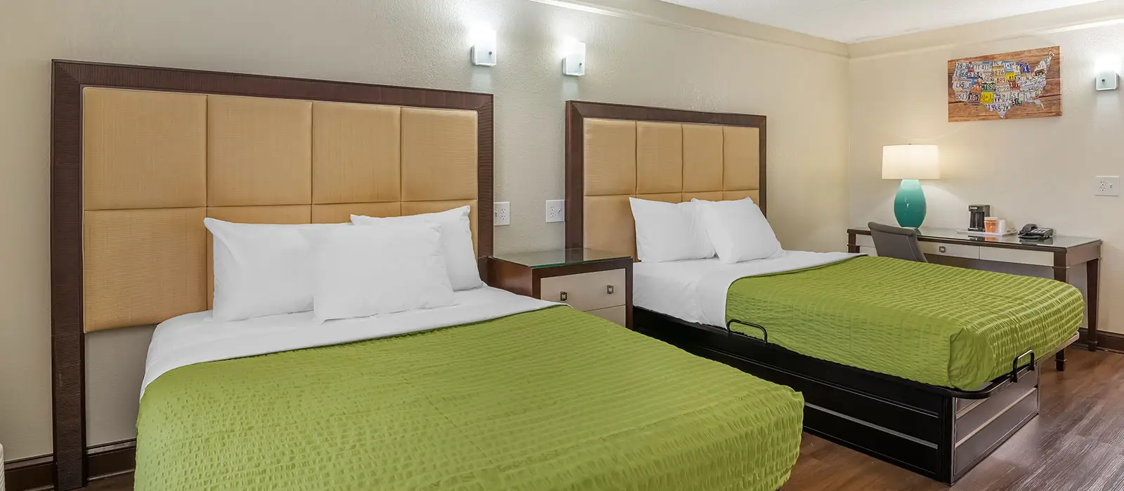 A guest room with two beds with green bedspreads, a bedside table between them and a desk with a lamp and a chair