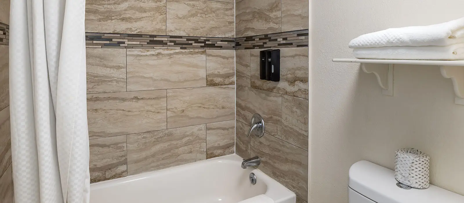 A bathroom with a white bathtub, a shower curtain, tiled walls, two wall-mounted soap dispensers, a toilet, and a shelf with folded towels