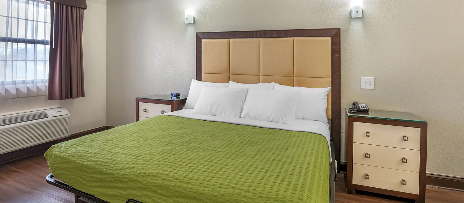 A guest room with a bed with a green bedspread, two bedside tables, and a window with sheer and brown curtains