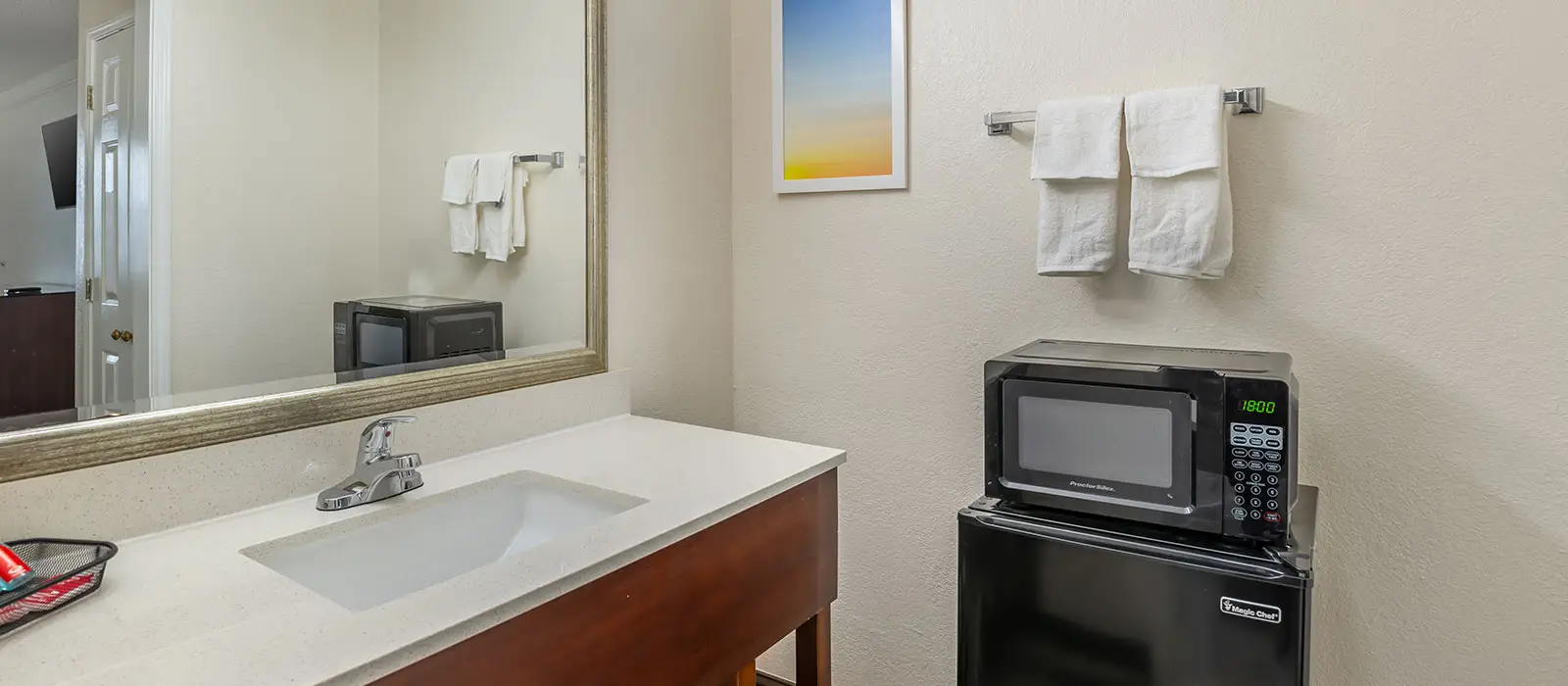 A bathroom vanity with a sink and faucet, a mirror, and a microwave and mini-fridge unit