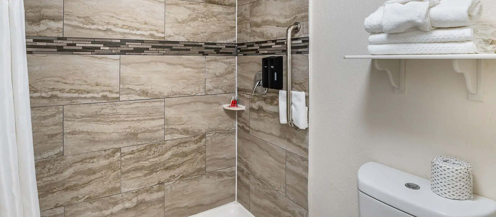 A tiled shower with a grab bar and dispensers, a shelf with towels, and a toilet with a roll of toilet paper