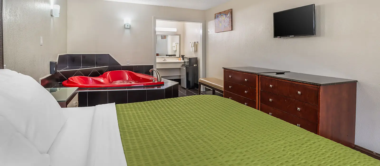 A guest room with a red heart-shaped jacuzzi tub, a green bedspread, a wall-mounted TV and a dresser