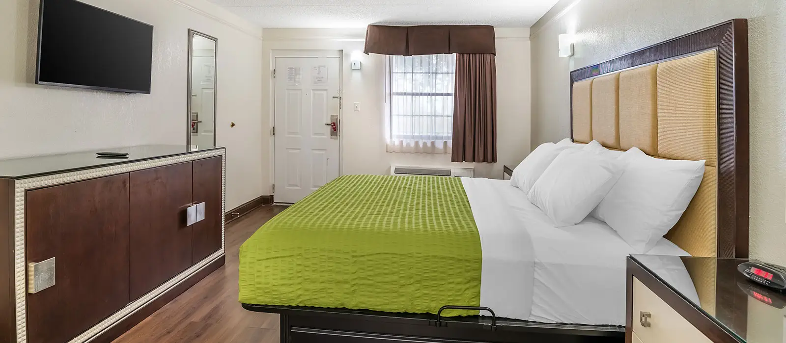 A guest room with a bed featuring a green bedspread, a wall-mounted TV above a long wooden cabinet, and a window with brown curtains