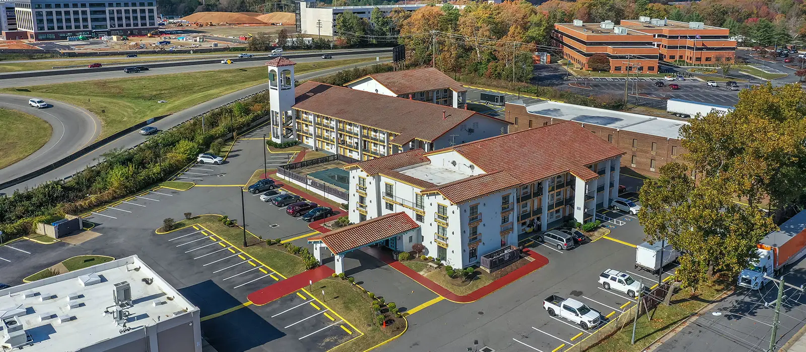 An aerial view of a hotel with a red tile roof, a swimming pool, and a parking lot, next to a highway