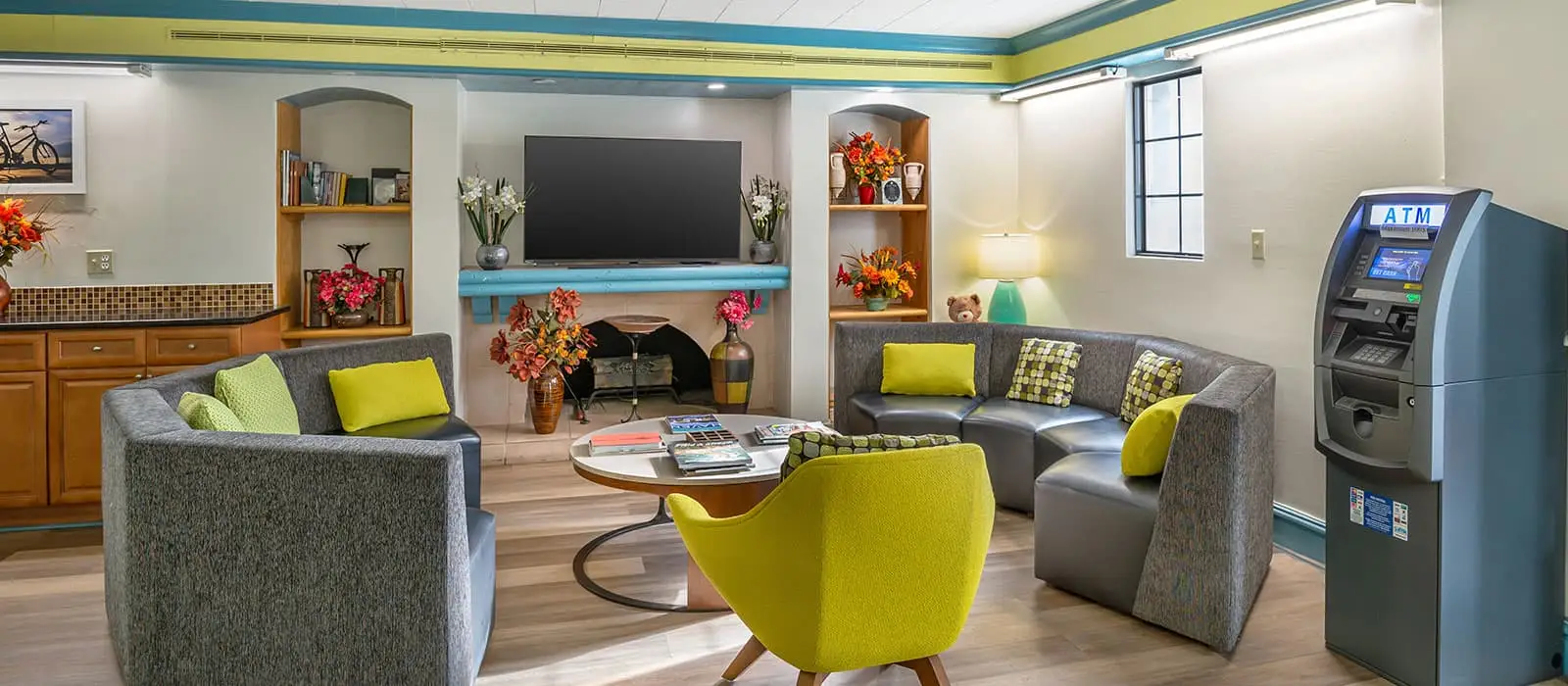 A hotel lobby with grey sectional sofas, a yellow armchair, a coffee table, a TV above a fireplace, and an ATM