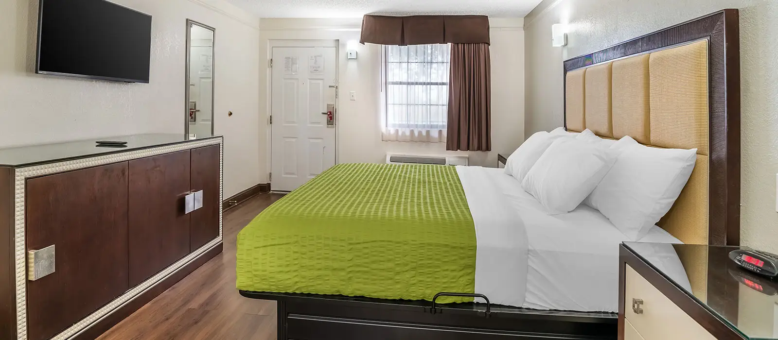 A hotel room with a bed featuring a green bedspread, a dark wood dresser with a TV, and a white door
